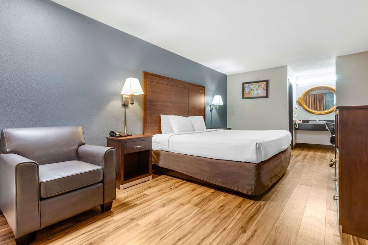A hotel room with a bed, a chair, a nightstand, a lamp, a painting, and a bathroom visible through an open door.