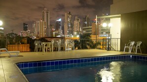 Una piscina en la azotea con vistas a la silueta urbana, rodeada de sillones de descanso y un área de estar cubierta.