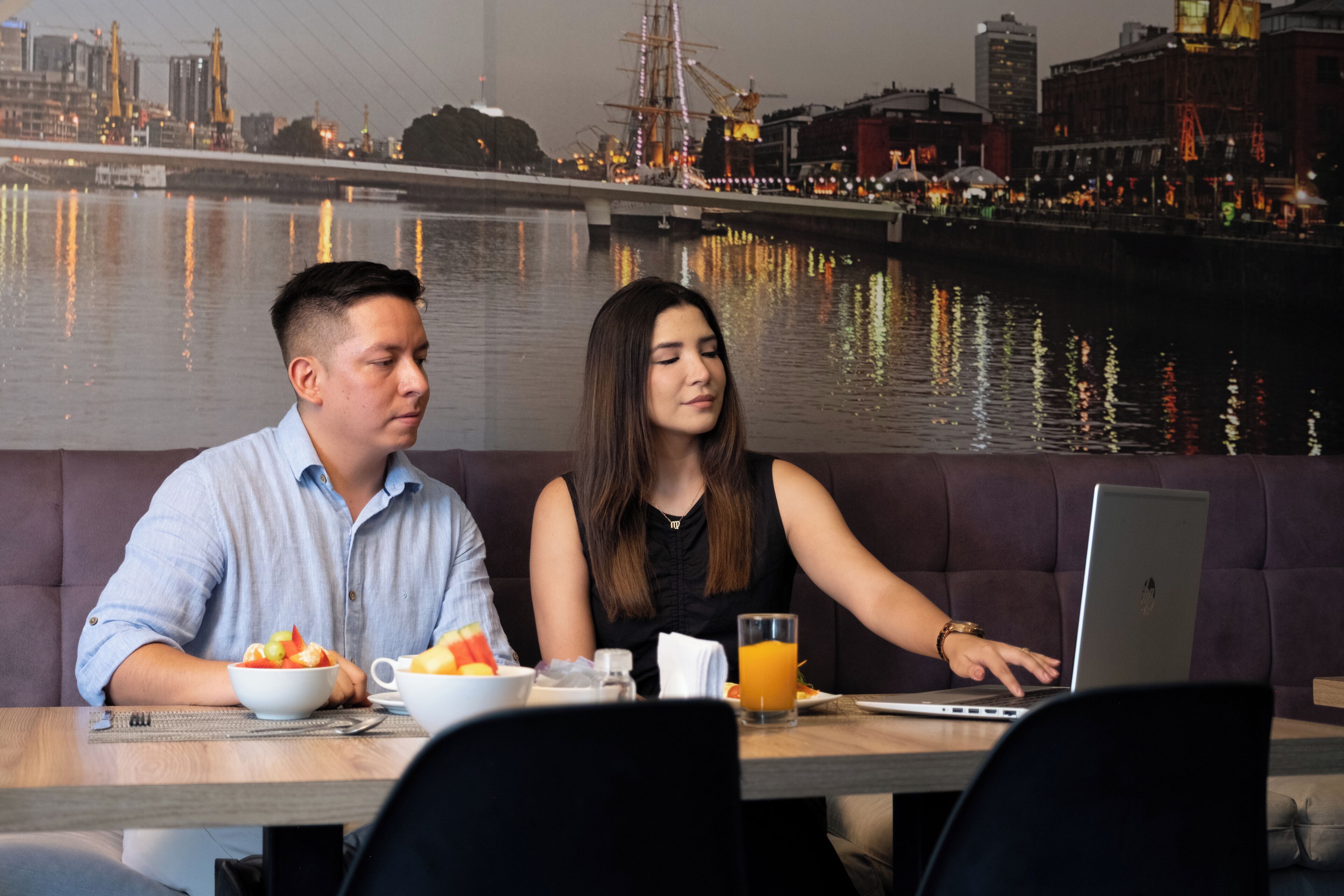 Two people seated at a table with a laptop, bowls of food, and a glass of orange juice. A cityscape with a waterfront is visible in the background.