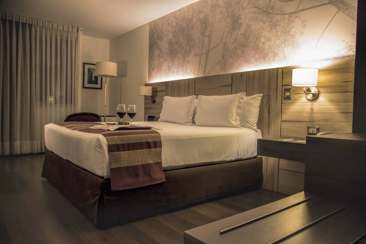 A modern hotel room with a large bed, bedside lamps, a wooden headboard, and a view of the outdoors through the window.
