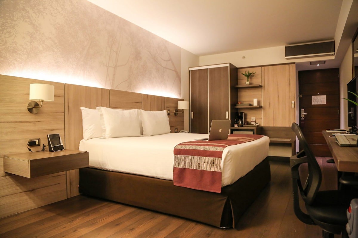 A hotel room with a bed, bedside table, lamp, laptop, desk, chair, and wardrobe.