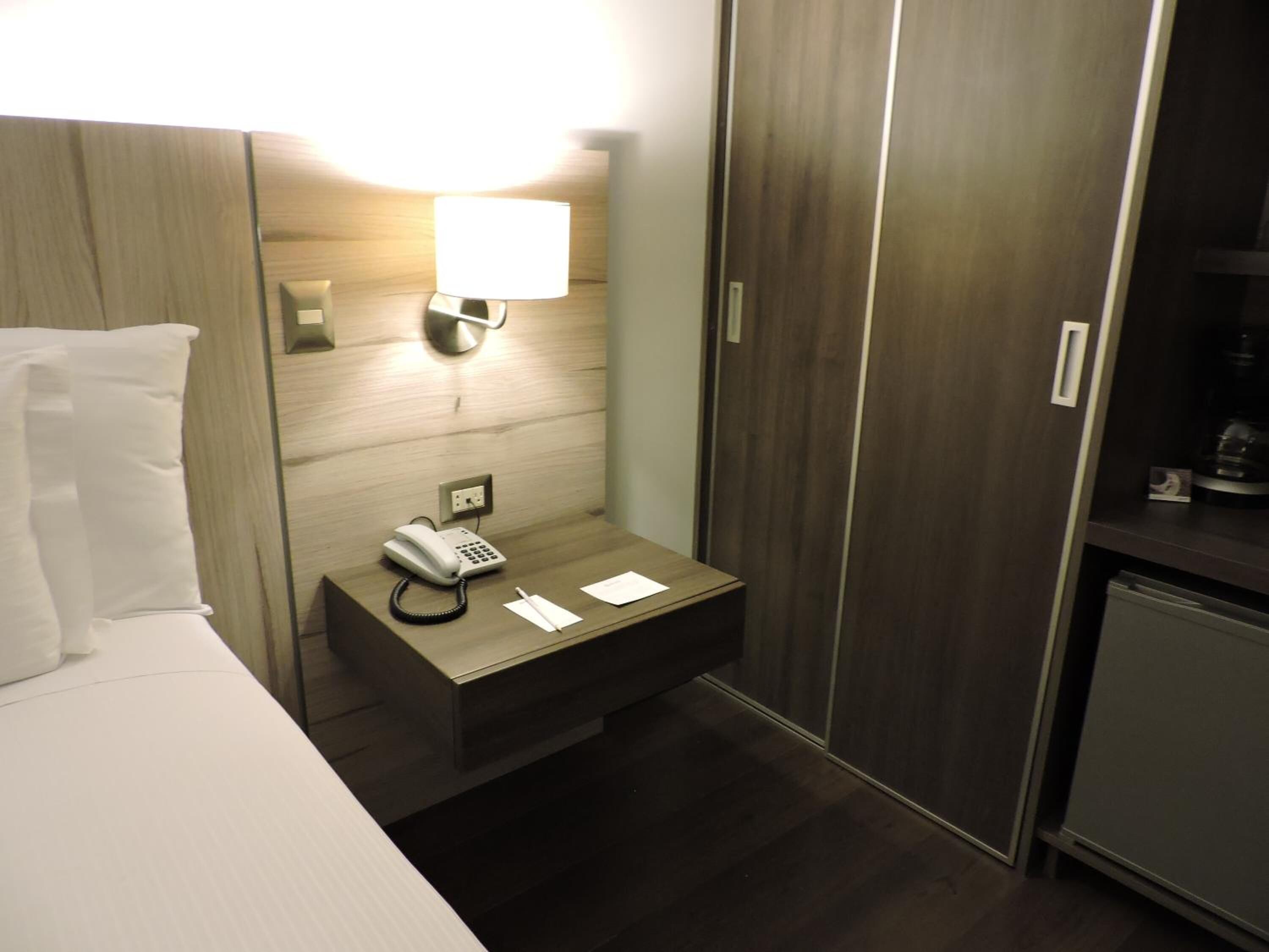 Et hotelværelse med en seng, et natbord med en telefon, en lampe og et klædeskab.