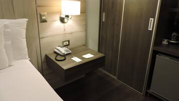 Habitación de hotel con cama, mesita de noche con teléfono, lámpara y armario.