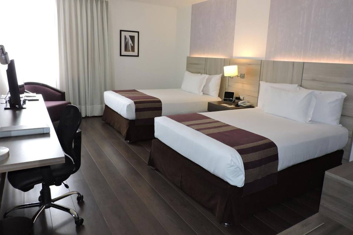 A hotel room with two beds, a desk, a chair, a lamp, and a picture on the wall.