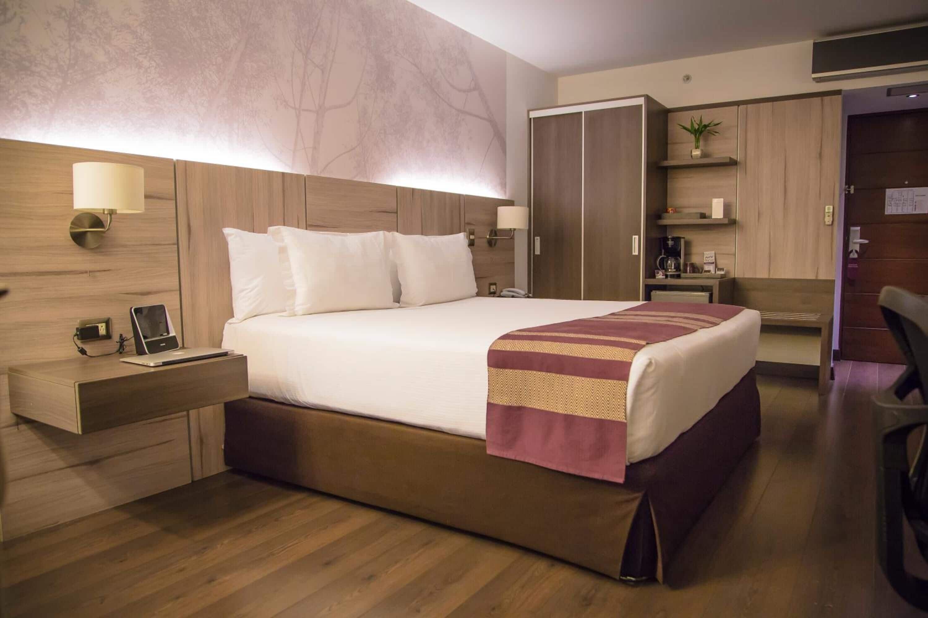 A hotel room with a large bed, wooden headboard, bedside lamps, a desk with a lamp, a coffee machine, and a wardrobe.