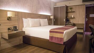 A hotel room with a large bed, wooden headboard, bedside lamps, a desk with a lamp, a coffee machine, and a wardrobe.