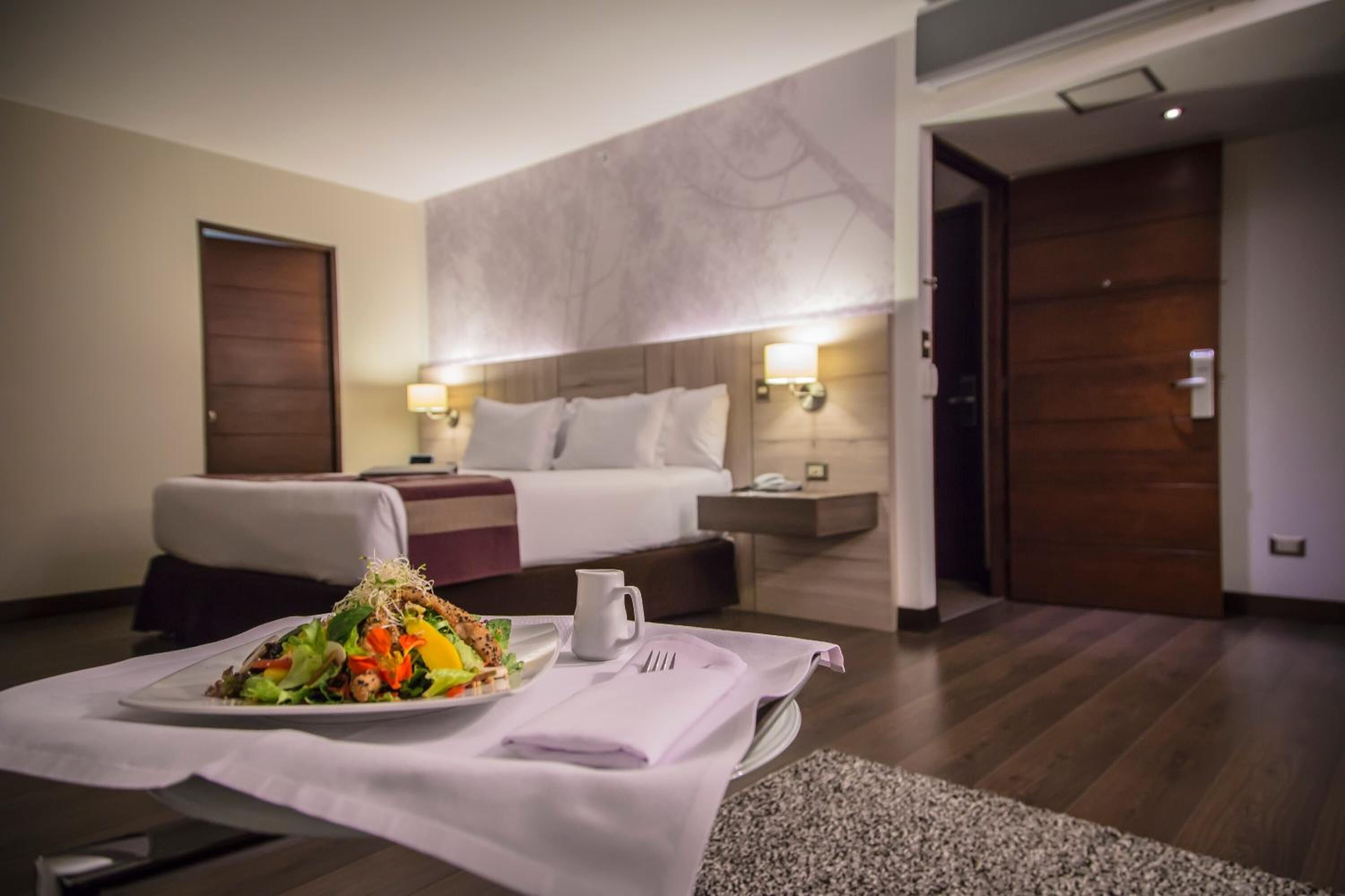 Una habitación de hotel con una cama bien hecha, una mesa de comedor con una ensalada y una mesita con una taza blanca.