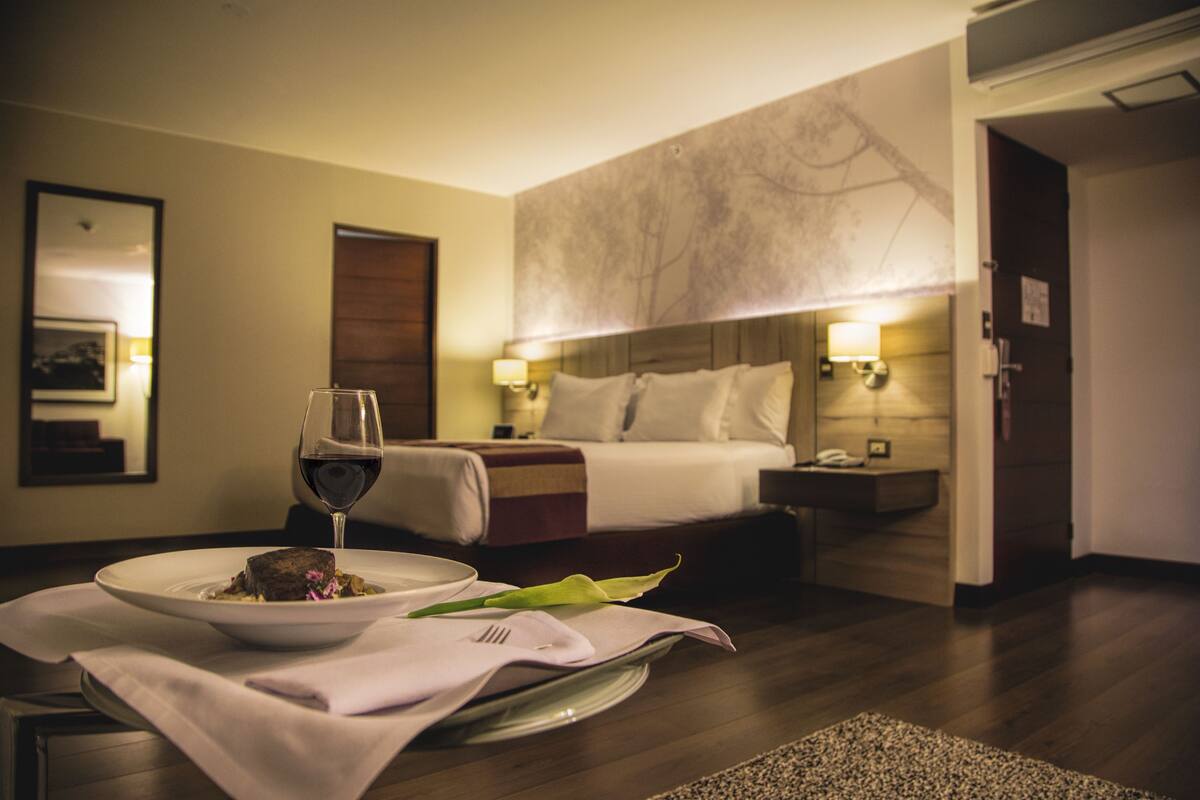 A hotel room with a bed, bedside tables, a dining table with a plate of food and a glass of wine, and a large abstract wall mural.