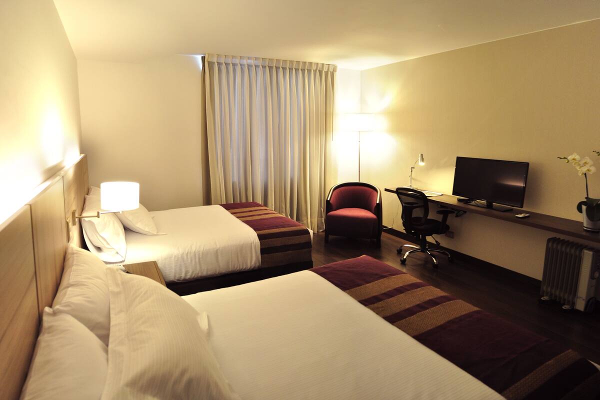 A hotel room with two beds, a desk with a computer, a chair, a TV, and a lamp.