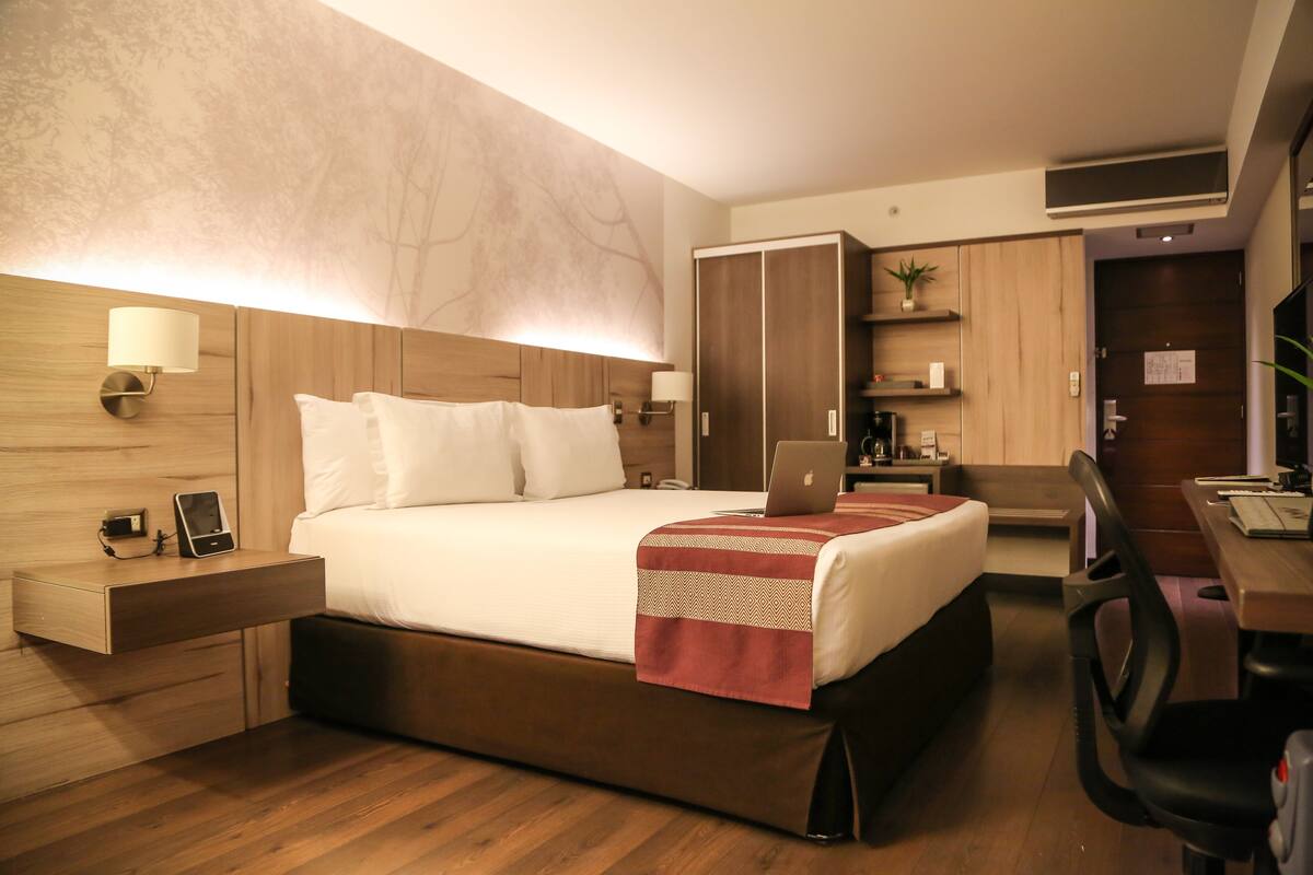 A modern hotel room with a wooden bed, bedside tables, a desk with a chair, and a wardrobe.
