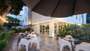 An outdoor patio area with white plastic chairs and tables, a large umbrella, and string lights. - Hotel MS Ciudad Jardín Plus (Cali)