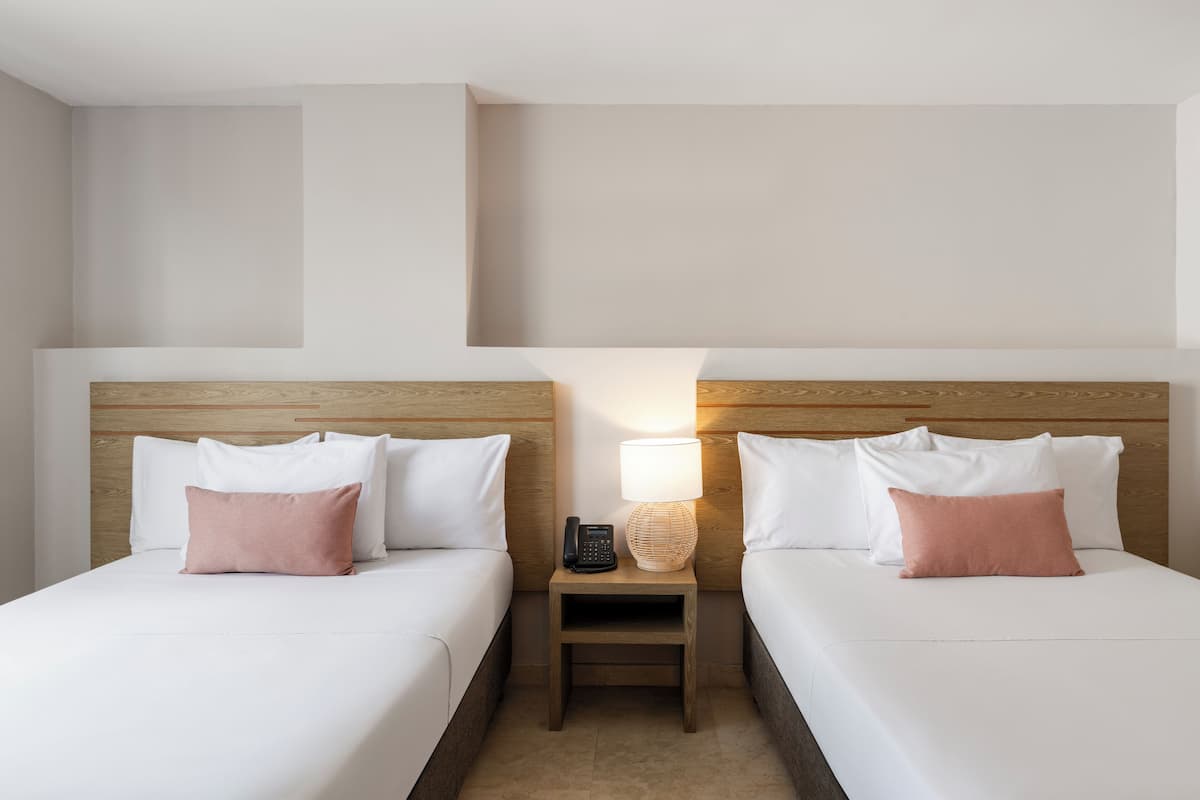 A hotel room with two beds, each with white bedding and a single pink pillow. There is a nightstand between the beds with a lamp and a telephone.