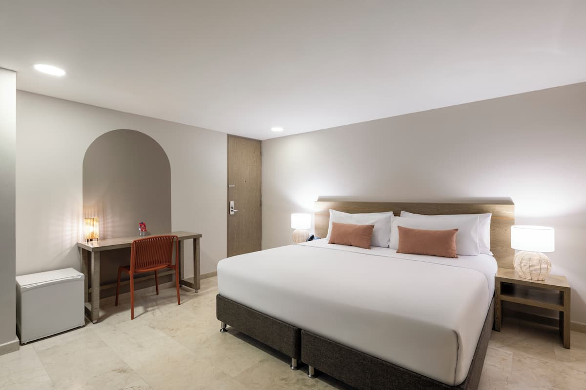 A hotel room with a large bed, a desk, a chair, a nightstand, and a small refrigerator.