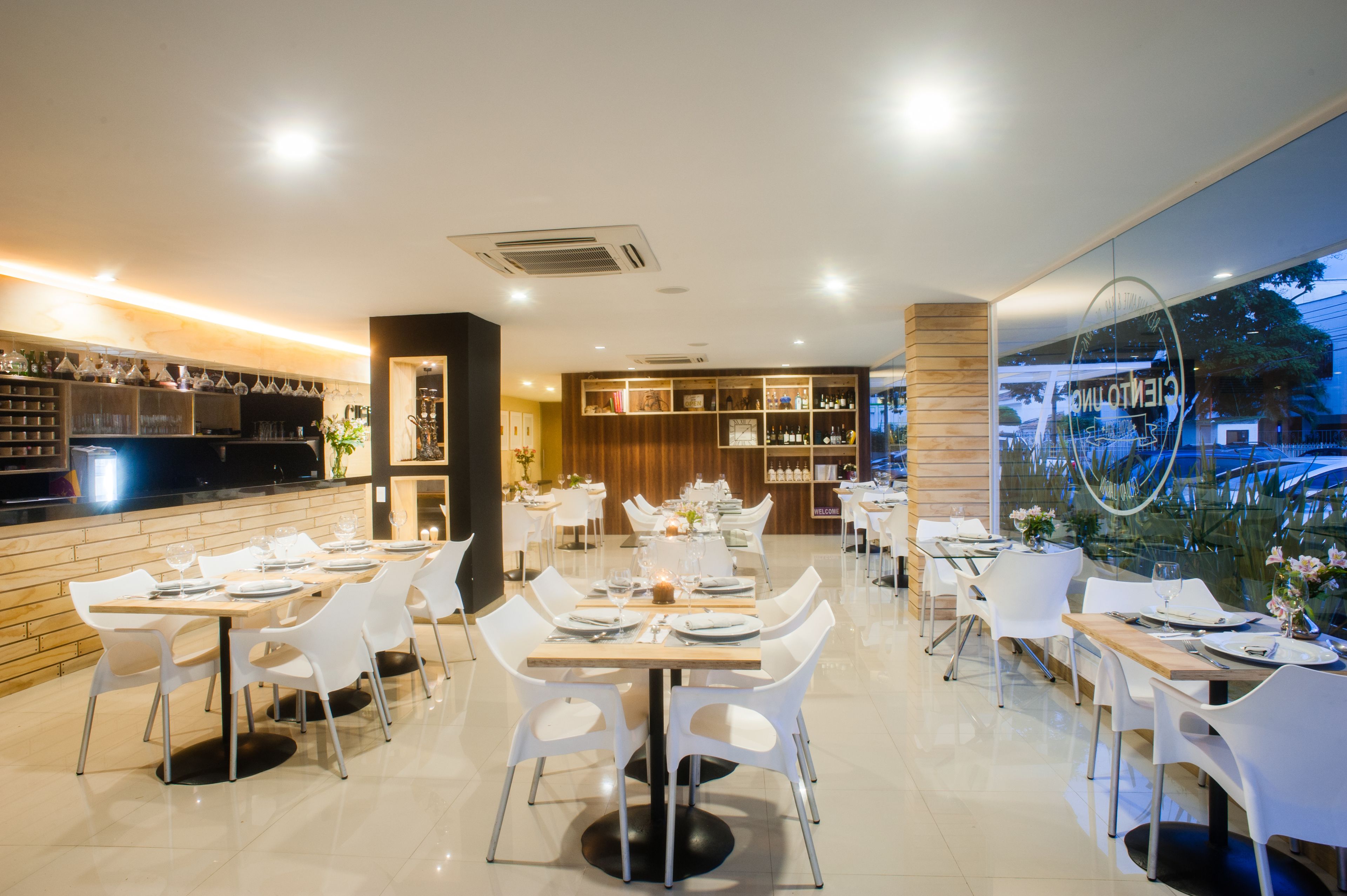 A modern restaurant with white chairs and tables, a bar area, and a view through a large window.