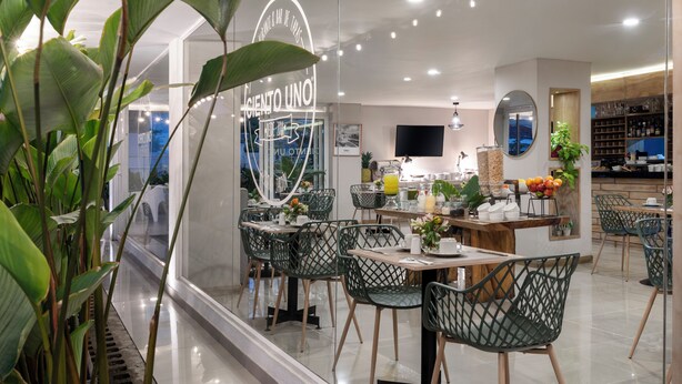 A modern cafe with a glass wall, green chairs, and a variety of plants. - Hotel MS Ciudad Jardín Plus (Cali)