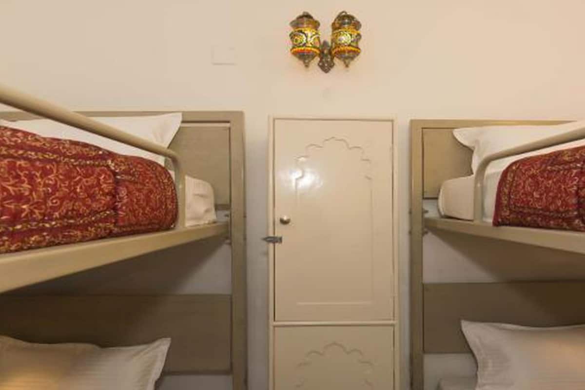 Une chambre avec deux lits superposés, chacun agrémenté d’une couvre-lit à motifs et de coussins blancs. On y trouve une porte ornée d’un panneau décoratif et un luminaire fixé au mur, juste au-dessus.