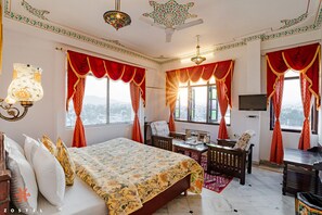 Une chambre d’hôtel spacieuse, dotée d’un grand lit, d’un bureau et d’une chaise. Elle comprend plusieurs fenêtres avec des rideaux rouges, un ventilateur de plafond et un téléviseur fixé au mur.