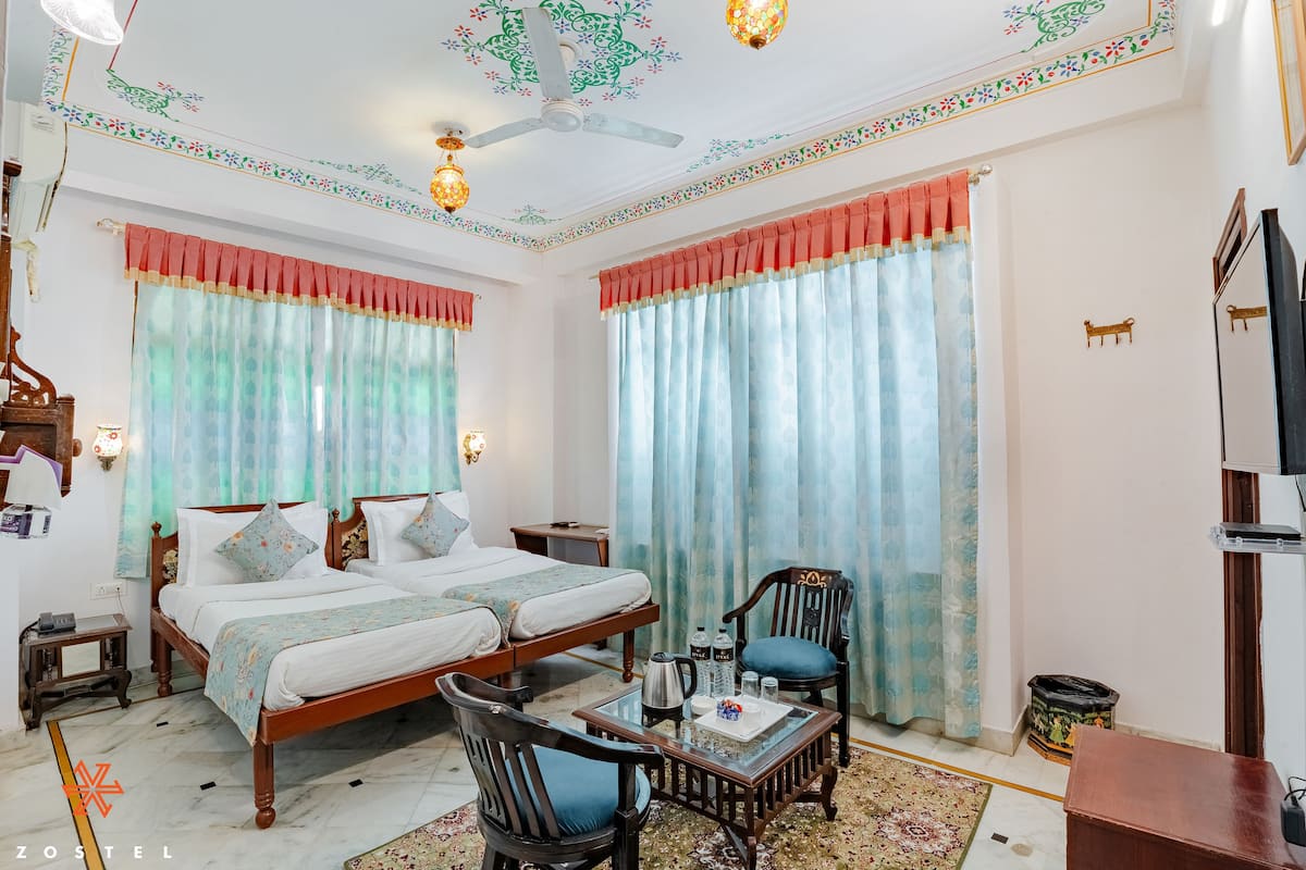 Une chambre d’hôtel avec deux lits, un coin salon avec une table et des chaises, un ventilateur de plafond, des luminaires décoratifs et un téléviseur.