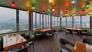 Un restaurante con varias mesas y sillas, ventanas amplias que dan a vistas de la ciudad, y coloridos faroles colgantes.