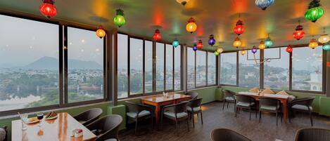 Un ristorante con diversi tavoli e sedie, ampie finestre che offrono vista sulla città e lanterne colorate appese.