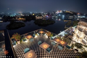 A rooftop terrace with checkered flooring, outdoor seating, and a view of a cityscape at night. - Zostel Udaipur - Hostel (Udaipur)