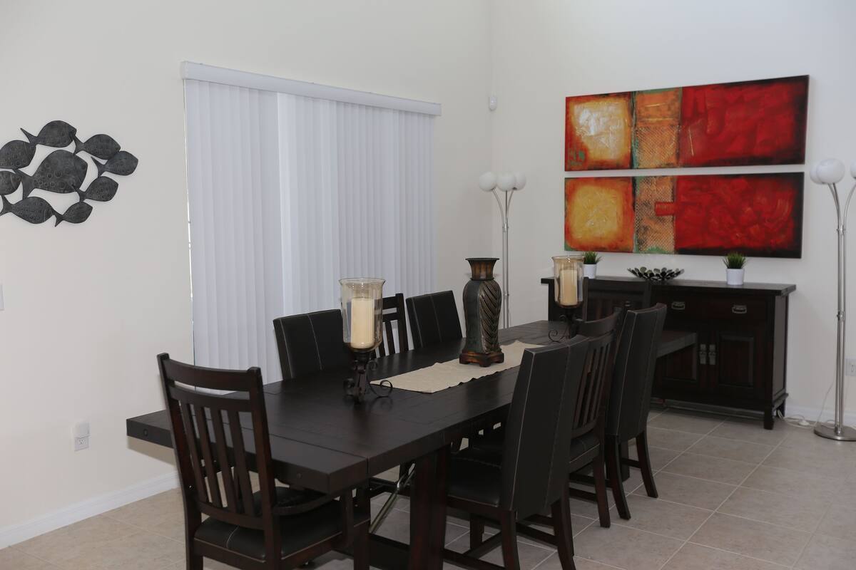 Uma sala de jantar com uma mesa de madeira escura, seis cadeiras, duas luminárias de chão altas e uma janela branca com persianas.