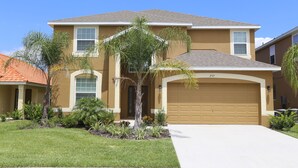 A two-story house with a beige exterior, a brown garage door, and a well-maintained front yard with various plants and a palm tree. - Orlando Select Vacation Homes (Kissimmee)