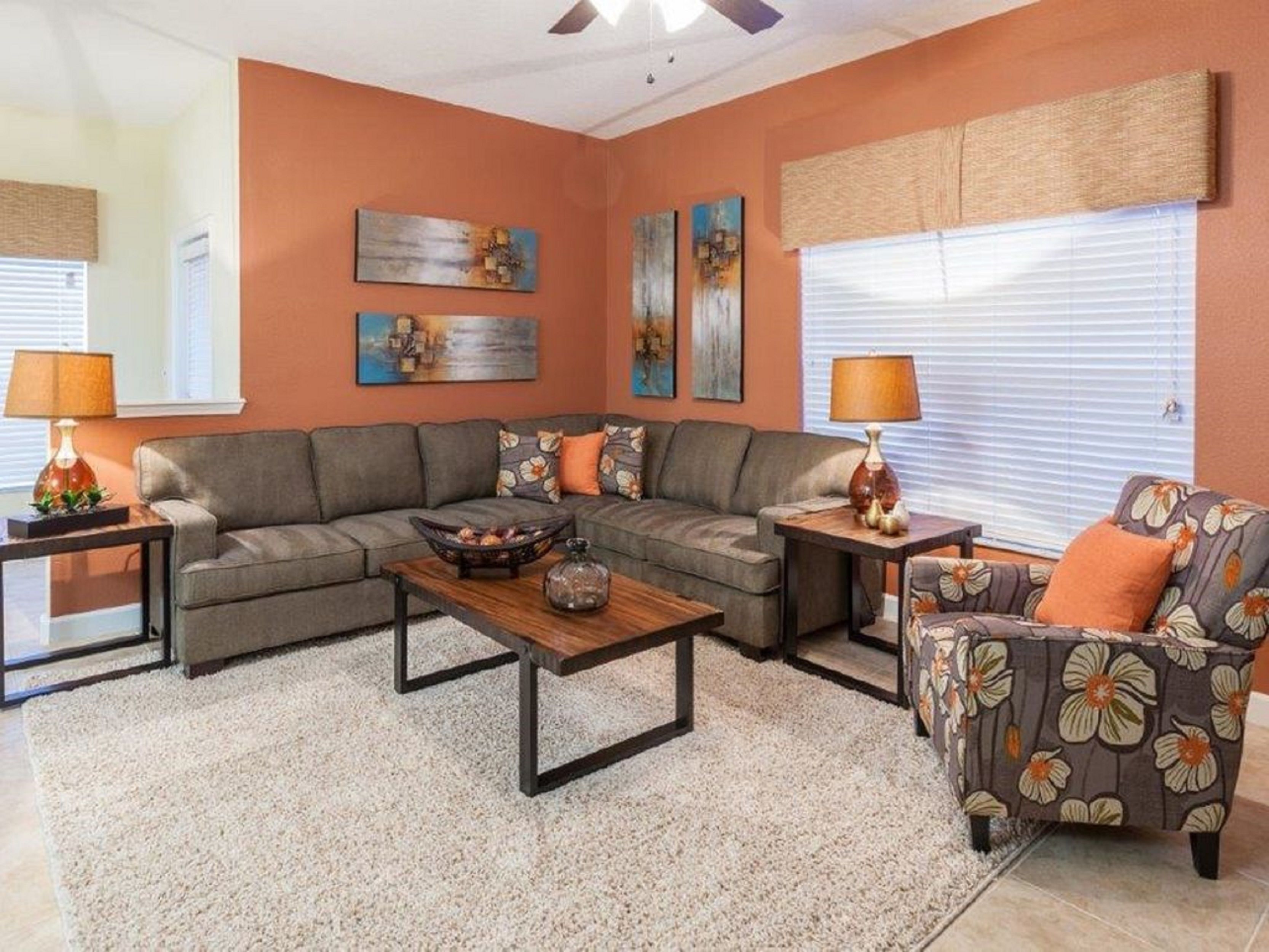 A living room with a sectional sofa, a coffee table, a floral armchair, and two side tables with lamps. There are abstract paintings on the walls and a window with blinds.