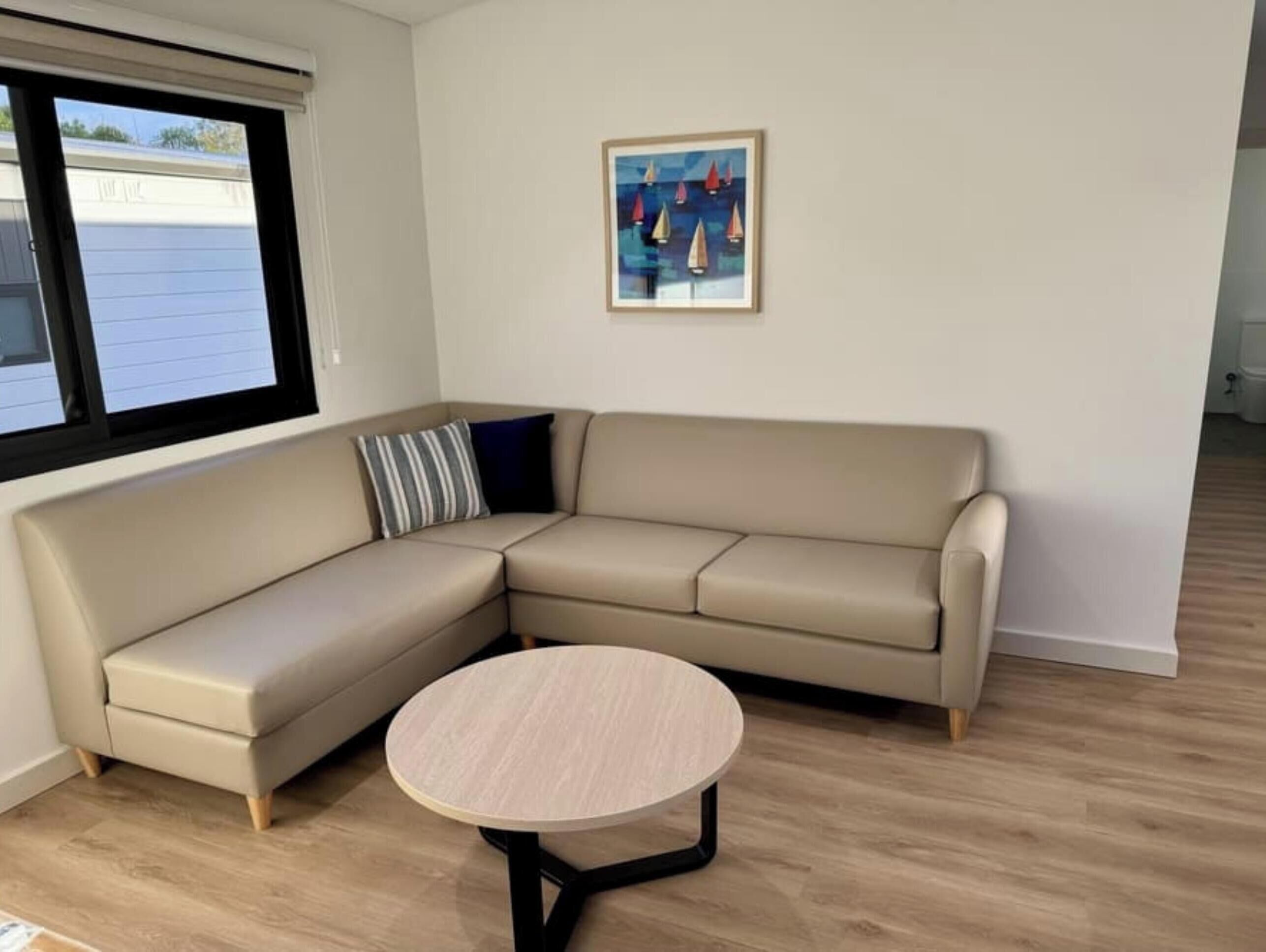 Una sala de estar en esquina con un sofá seccional color beige, una mesa redonda y un cuadro enmarcado con barcos a vela en la pared.