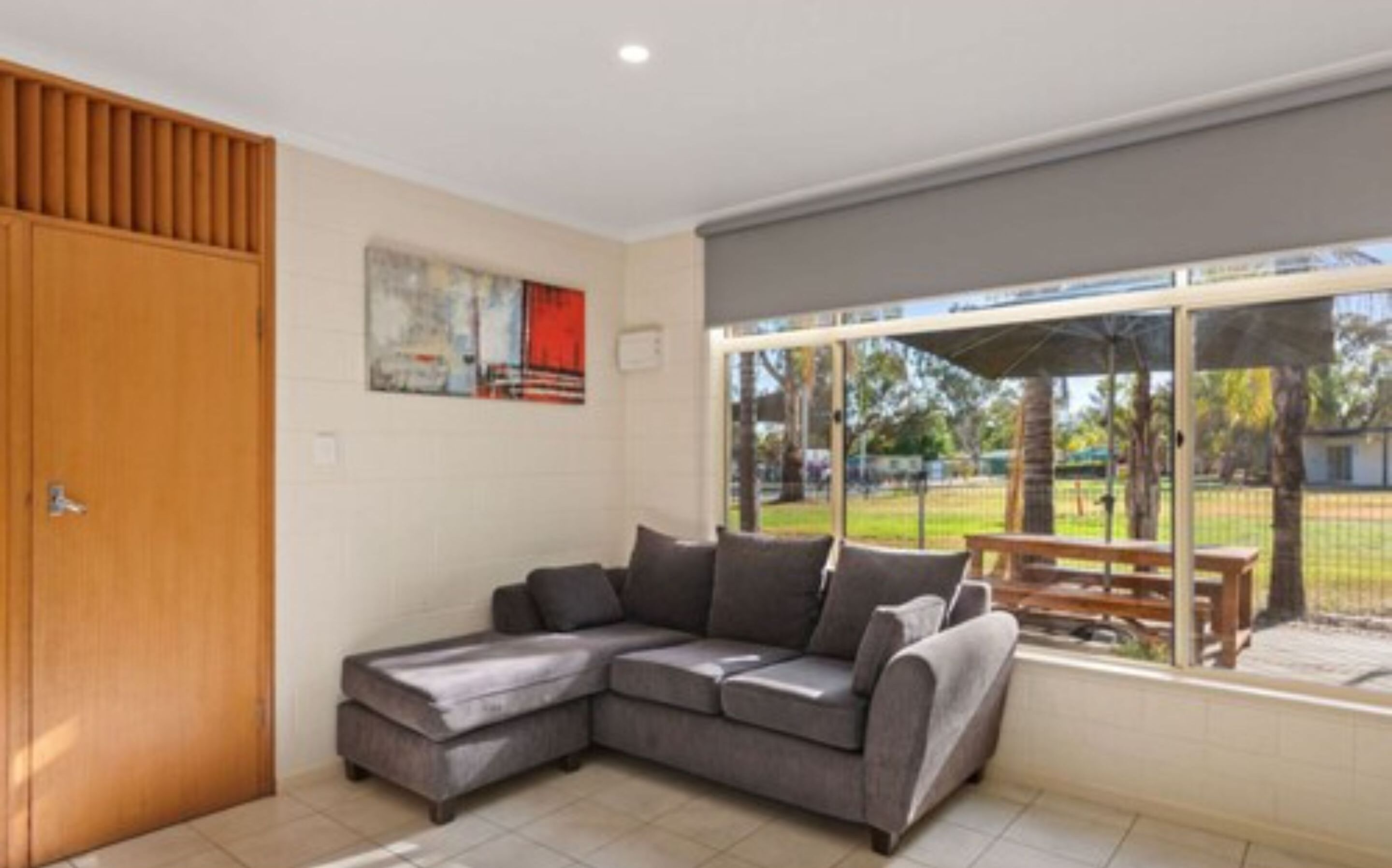 A modern living room with a grey sectional sofa, a wooden door, and a large window with a view of a park.