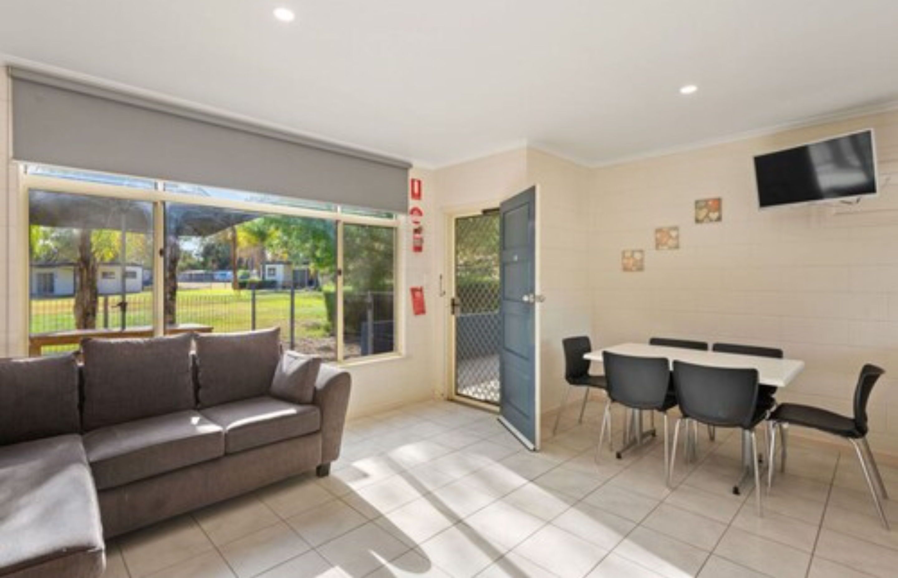 A modern room with a sofa, a dining table with chairs, a TV mounted on the wall, and a large window with a view of the outdoors.