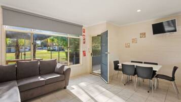 A modern room with a sofa, a dining table with chairs, a TV mounted on the wall, and a large window with a view of the outdoors.