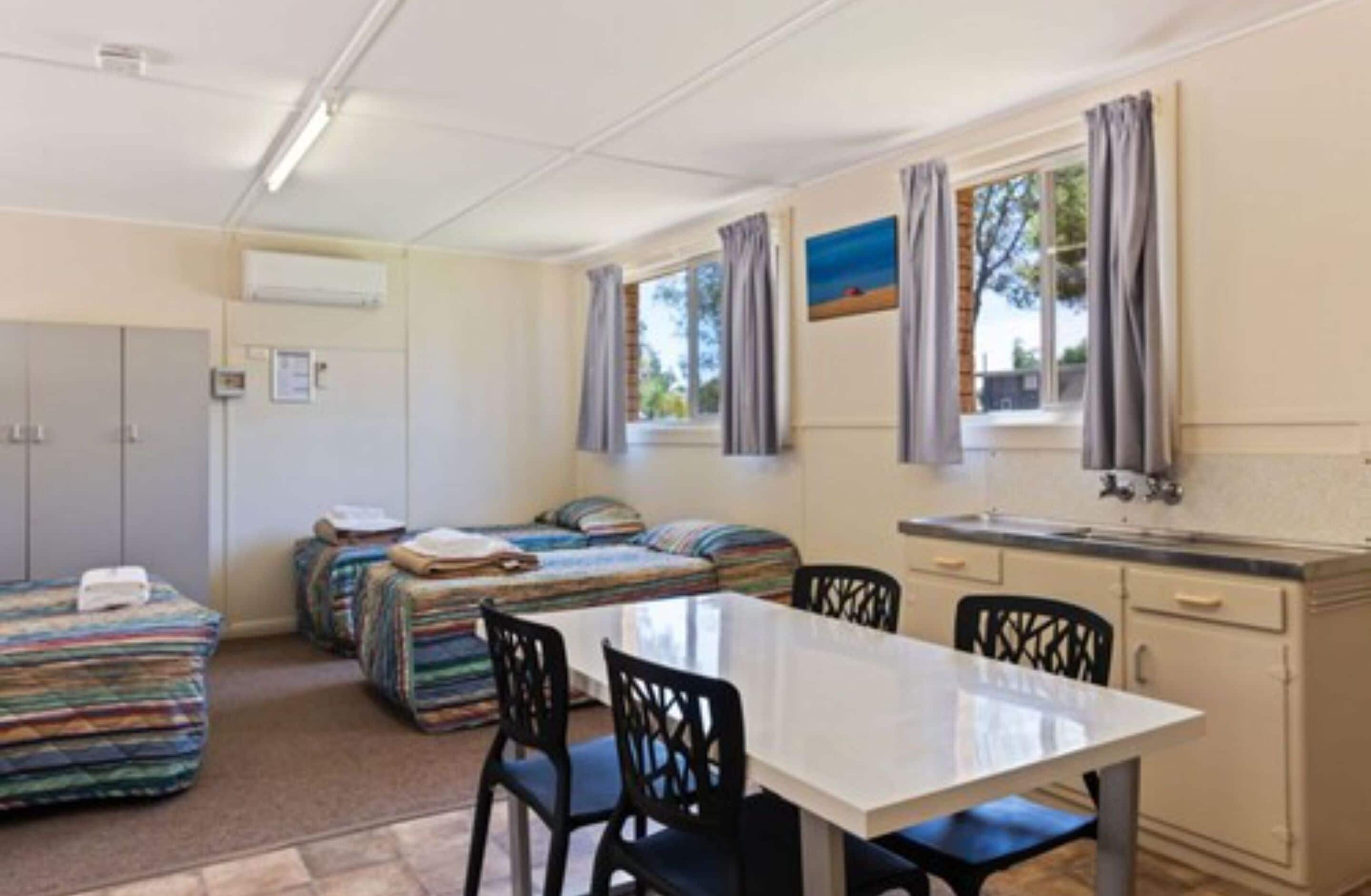 Une pièce compacte avec une kitchenette, deux lits dotés de draps colorés, une table à manger avec des chaises et une fenêtre avec des rideaux.
