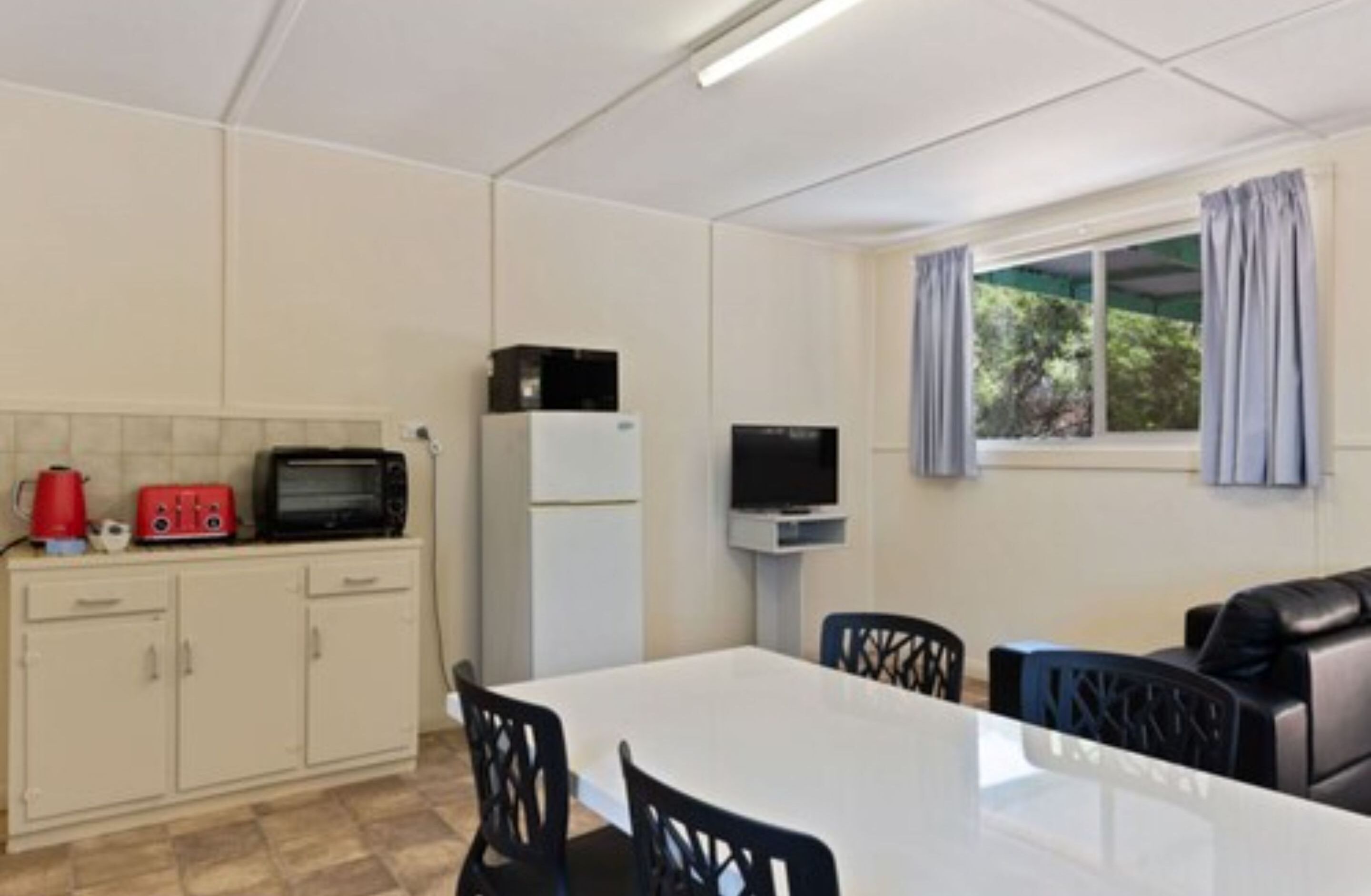 A compact kitchen with a microwave, toaster, and kettle, a dining area with a table and chairs, and a window with curtains.