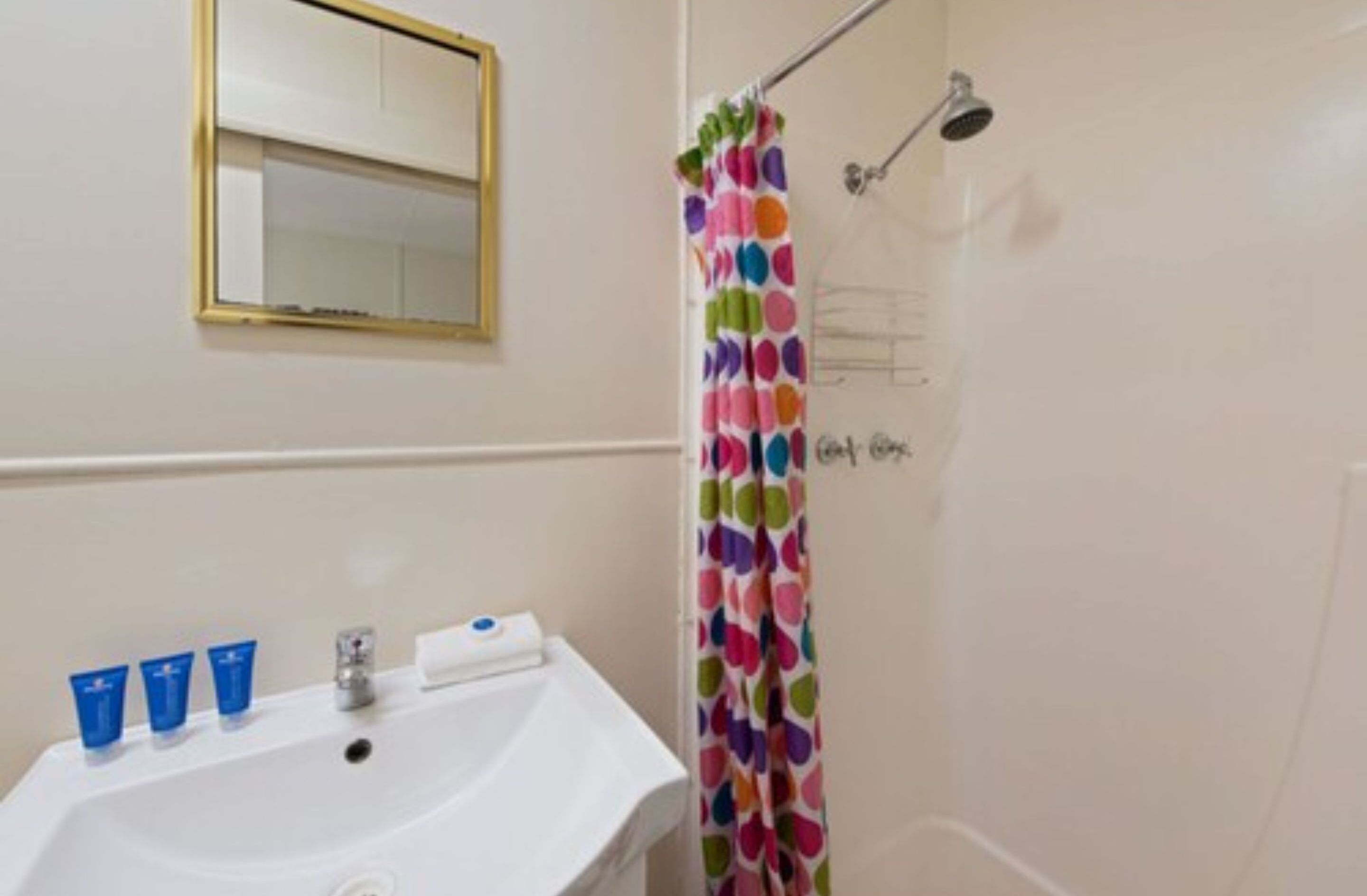 Un bany amb un lavabo blanc, tres productes de tocador blaus, un mirall amb marc daurat i una cortina de bany multicolor.