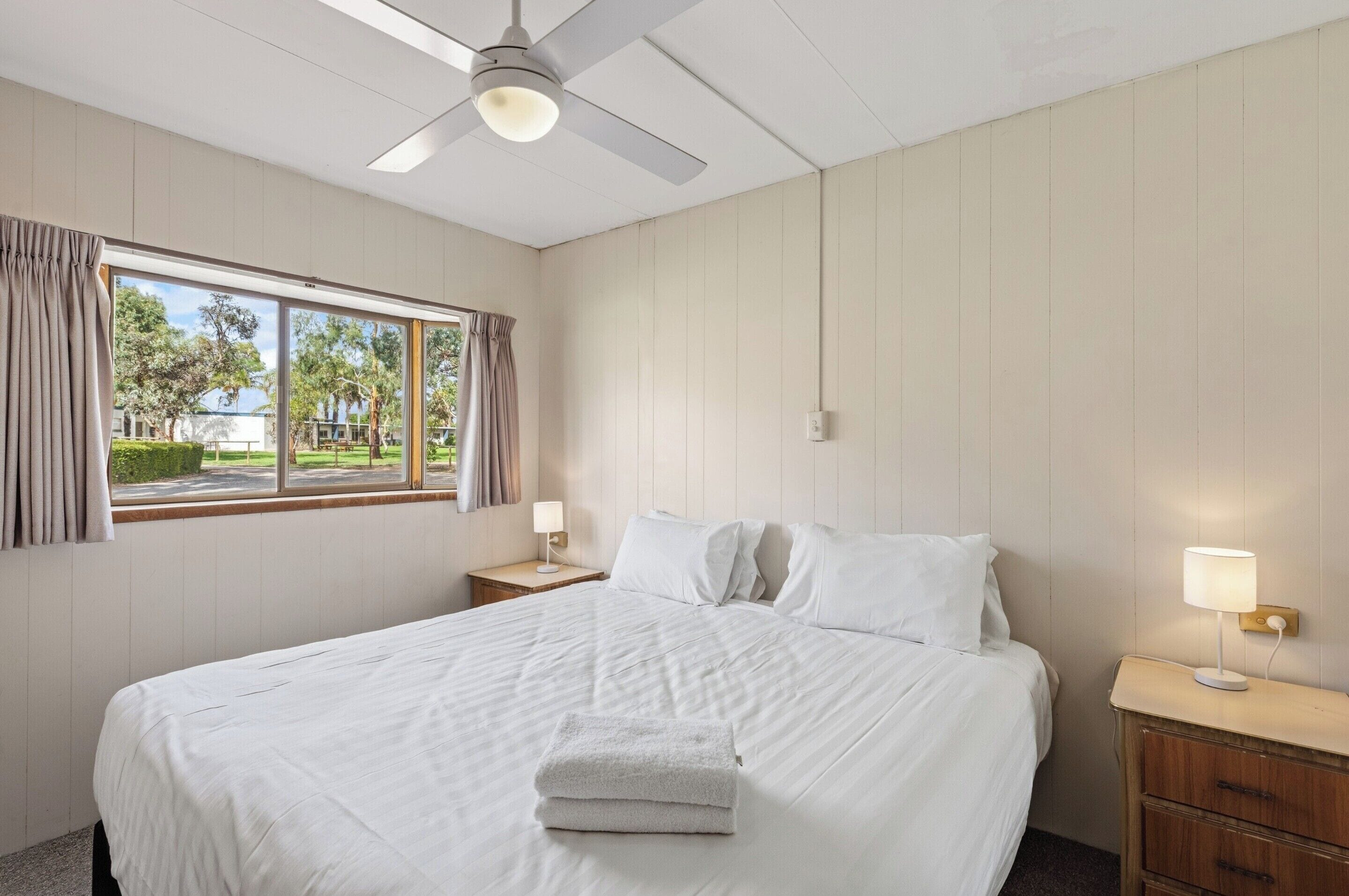 A hotel room with a bed, a nightstand, a lamp, a ceiling fan, and a window with curtains.