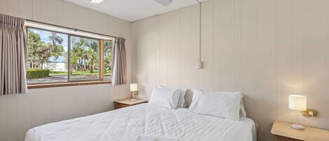 Ett hotellrum med en säng, ett nattduksbord, en lampa, en takfläkt och ett fönster med gardiner.