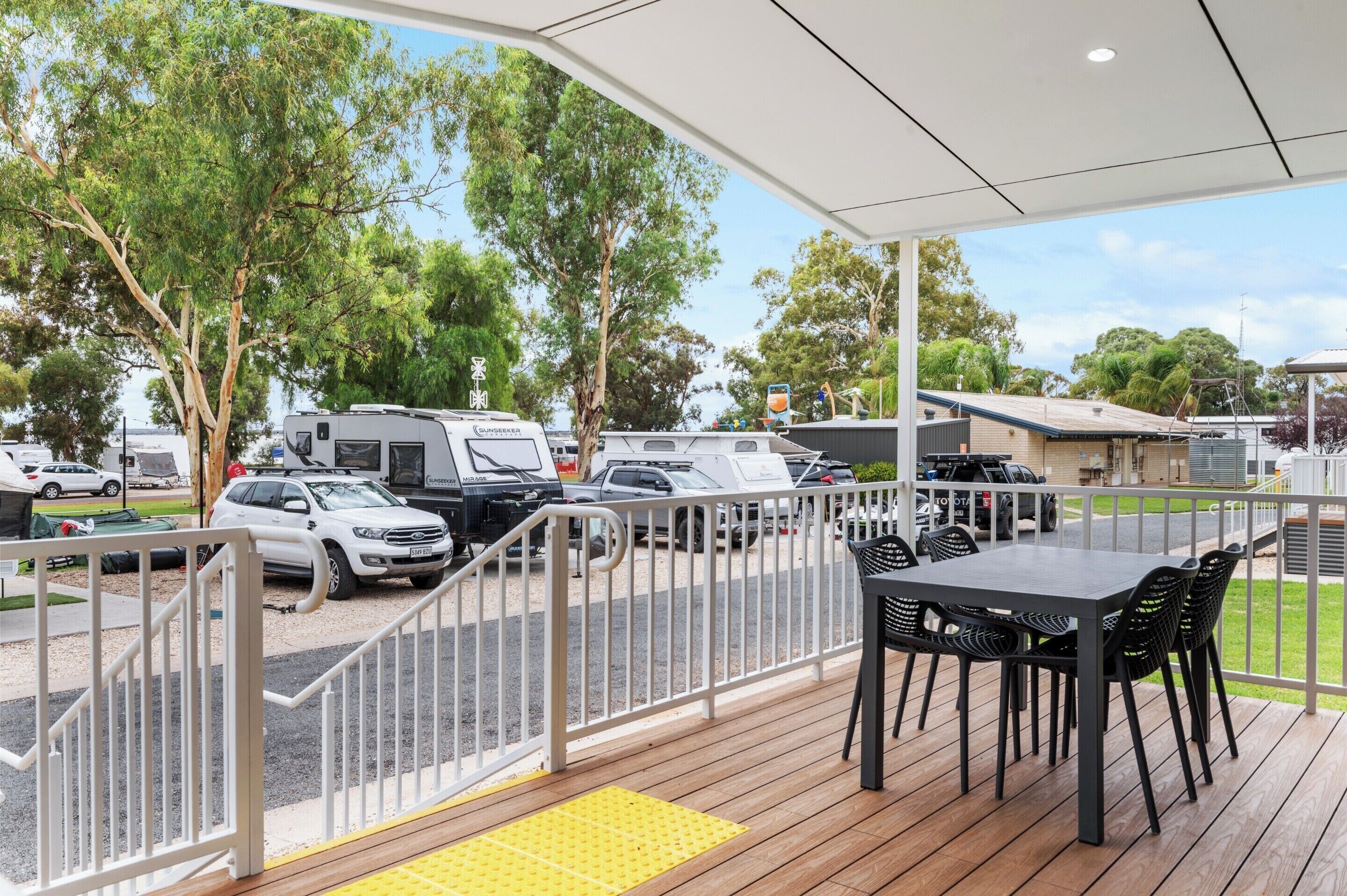 A covered outdoor area with a table and chairs, a white railing, and a view of parked vehicles and trees.