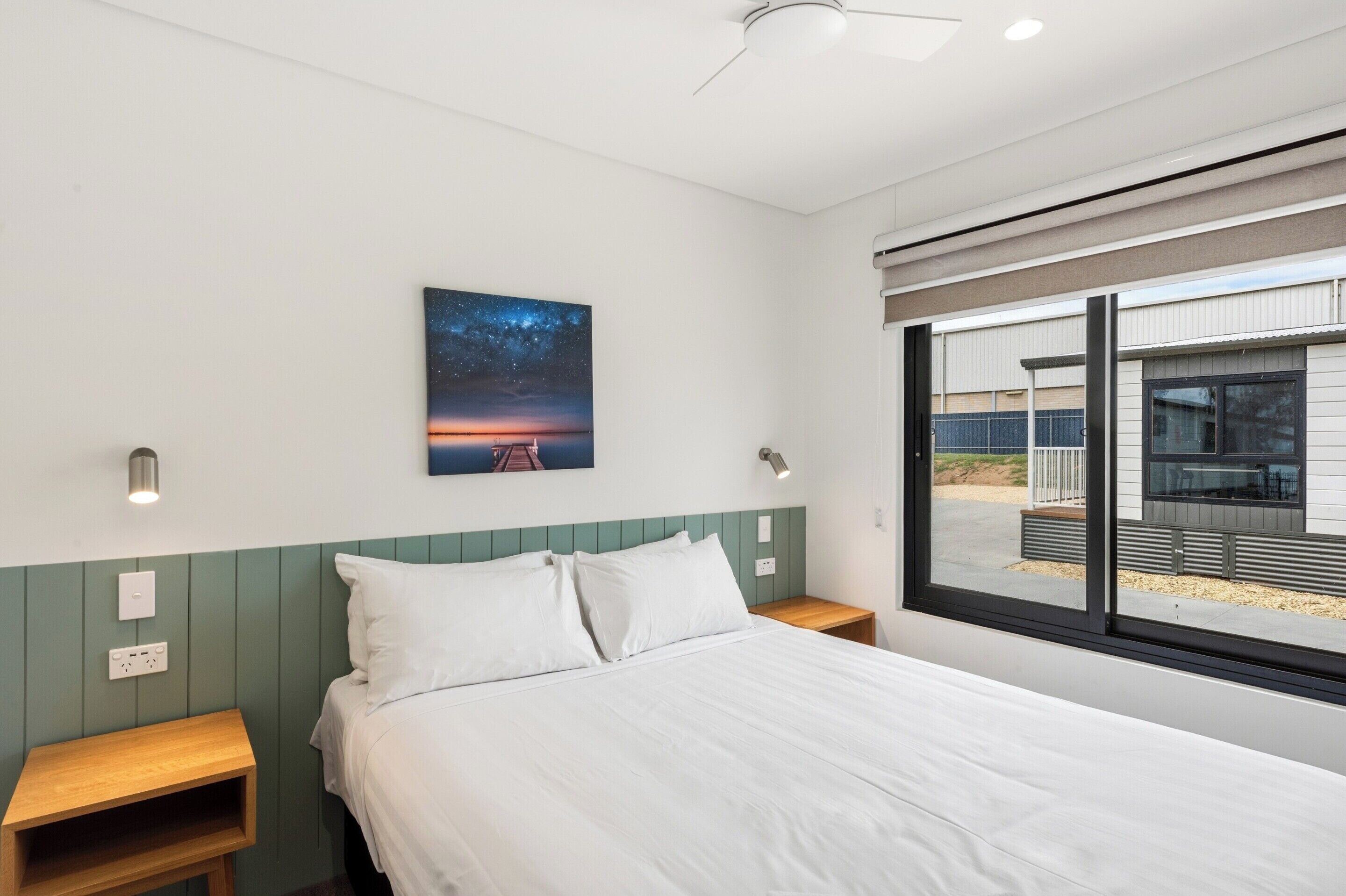 Un dormitorio con cama, mesitas de noche, una lámpara de pared, un cuadro y una ventana con vista a un edificio.