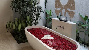 A bathtub filled with red and white rose petals, surrounded by lush green plants and a decorative butterfly sculpture. - Nyaman Villas (Seminyak)