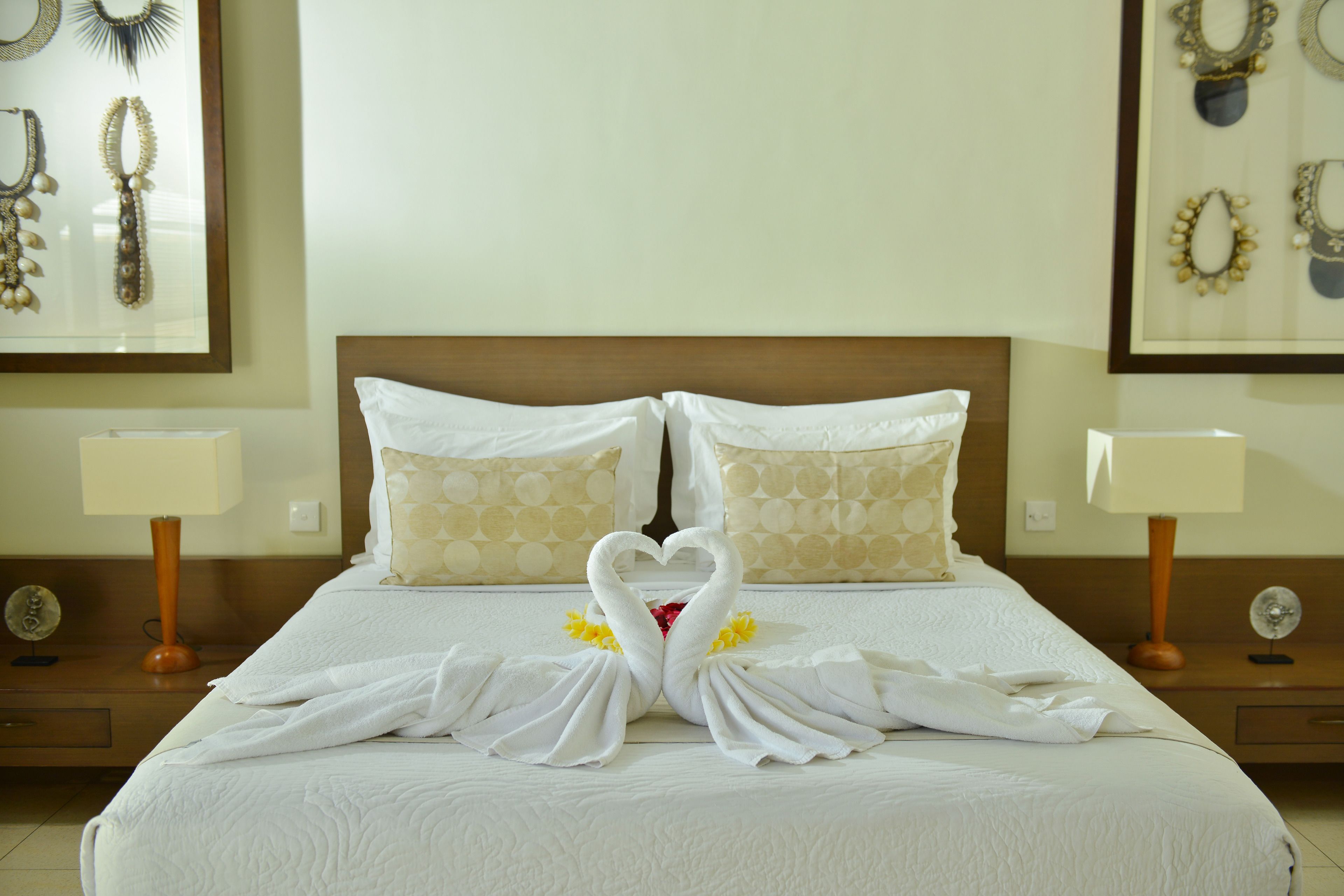 A hotel room with a neatly made bed, two pillows, a headboard, bedside tables with lamps, and framed jewelry on the walls.