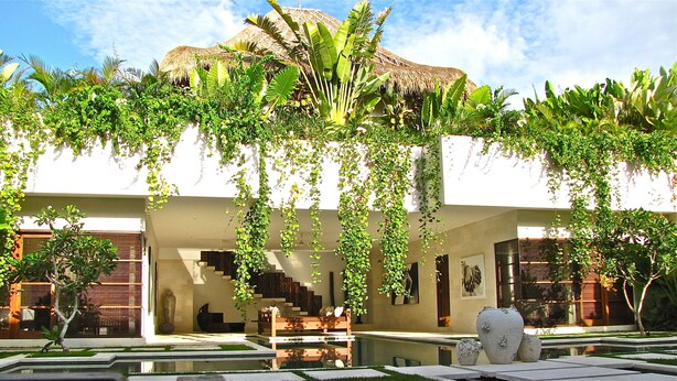 A modern building with a white exterior, a thatched roof structure, and lush greenery including hanging plants and trees. - Nyaman Villas (Seminyak)