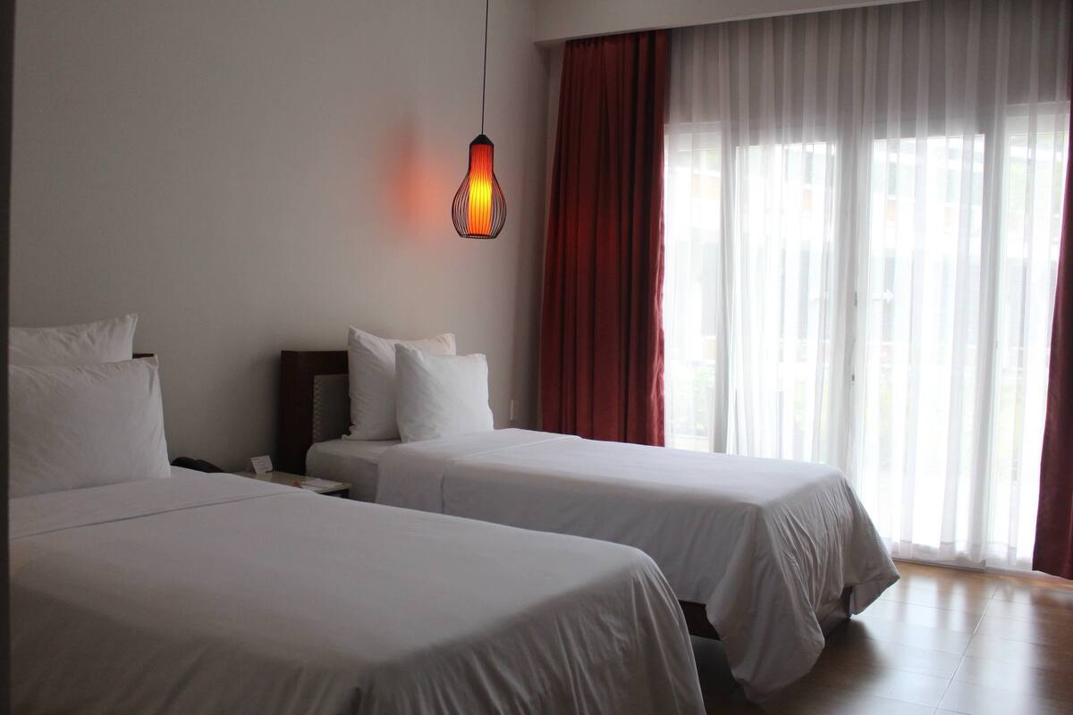 Una habitación de hotel con dos camas, una mesita de noche, una lámpara y una ventana con cortinas.