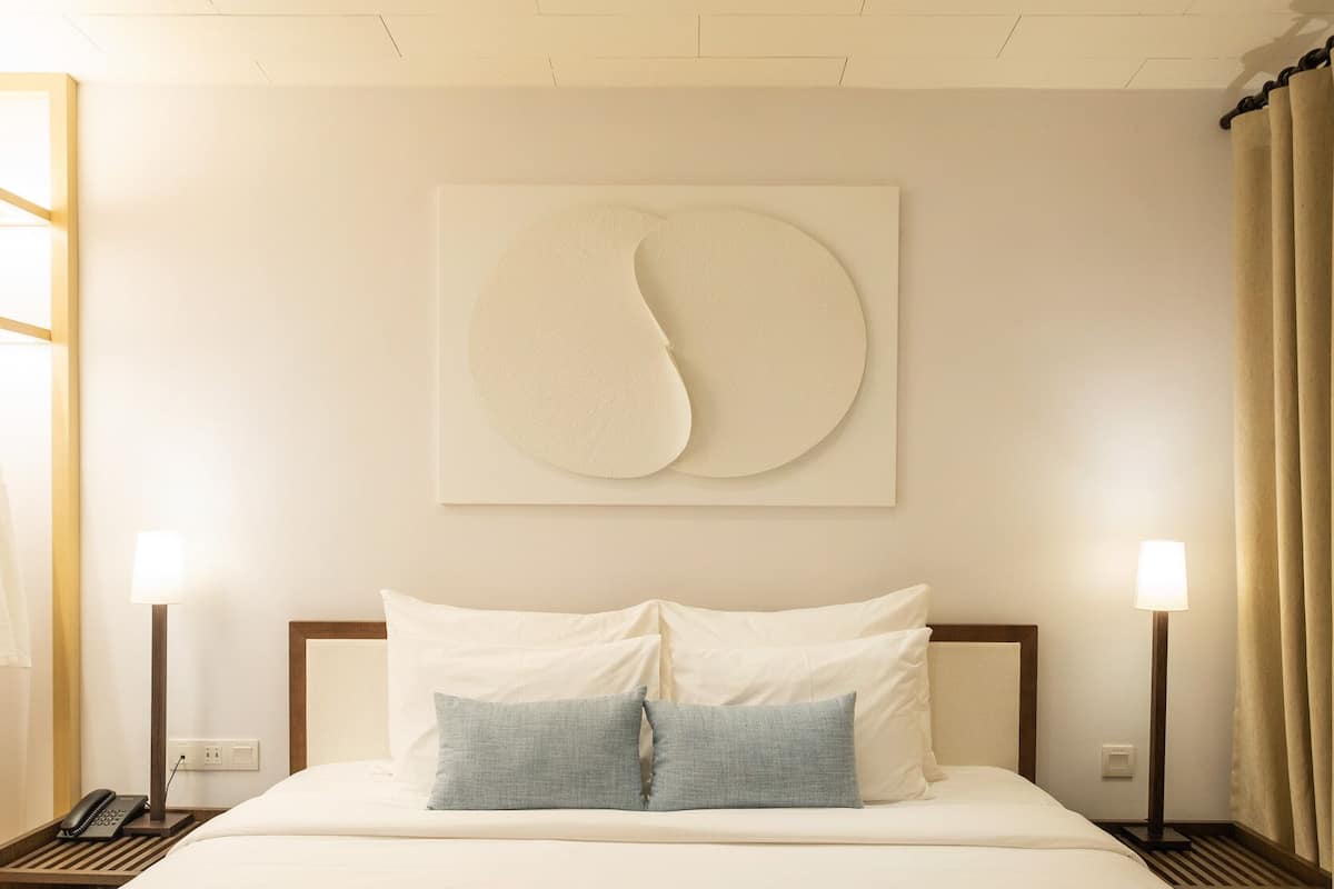 Habitación de hotel con una cama grande, dos mesitas de noche, un ventilador de techo y una obra de arte abstracta colgada en la pared.