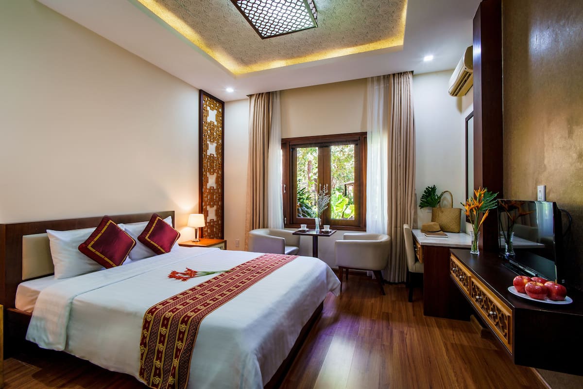 Una habitación de hotel con una cama grande, un escritorio con televisor, una mesita pequeña y una ventana con cortinas.