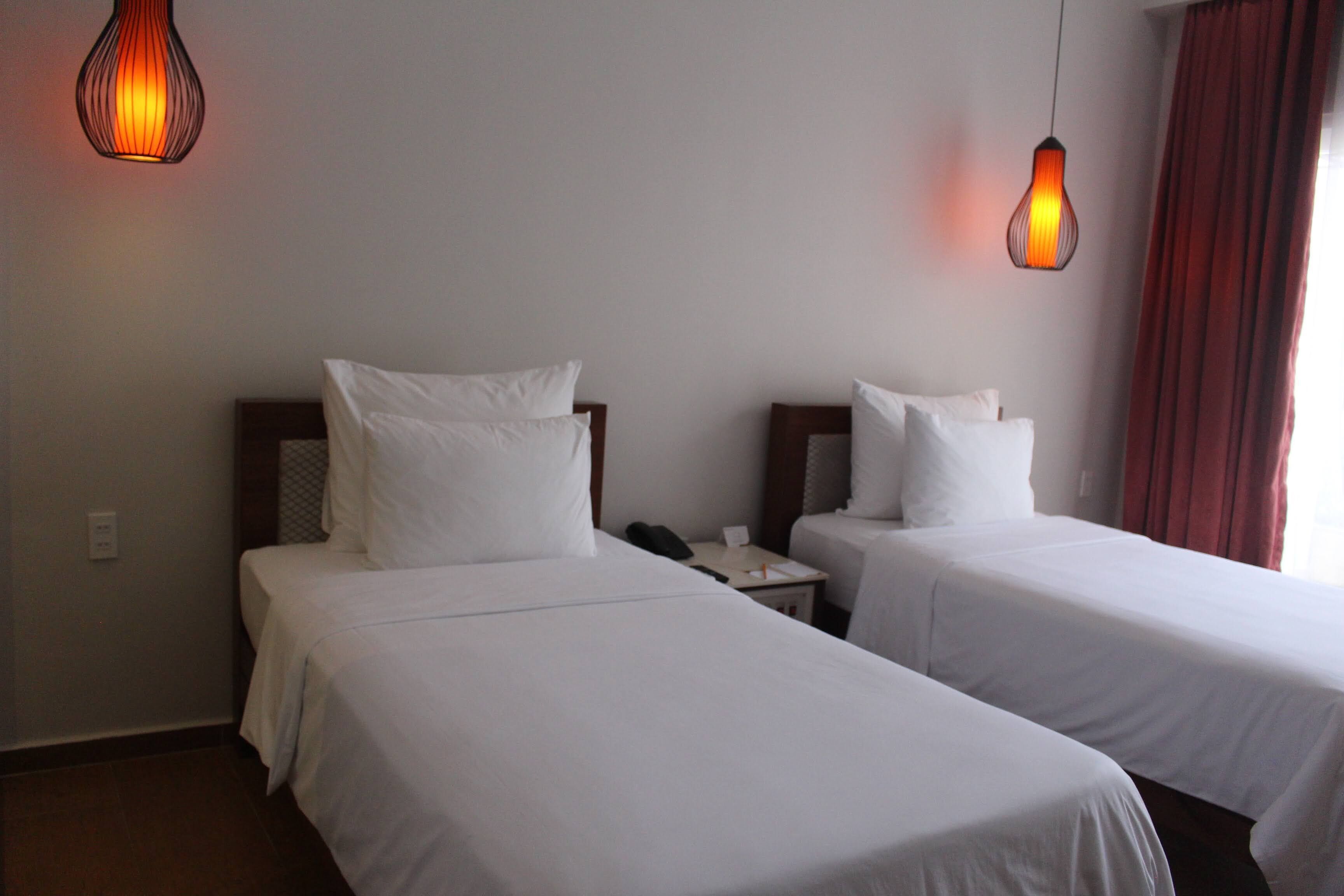 A hotel room with two single beds, a bedside table with a phone, and a wall-mounted light fixture.