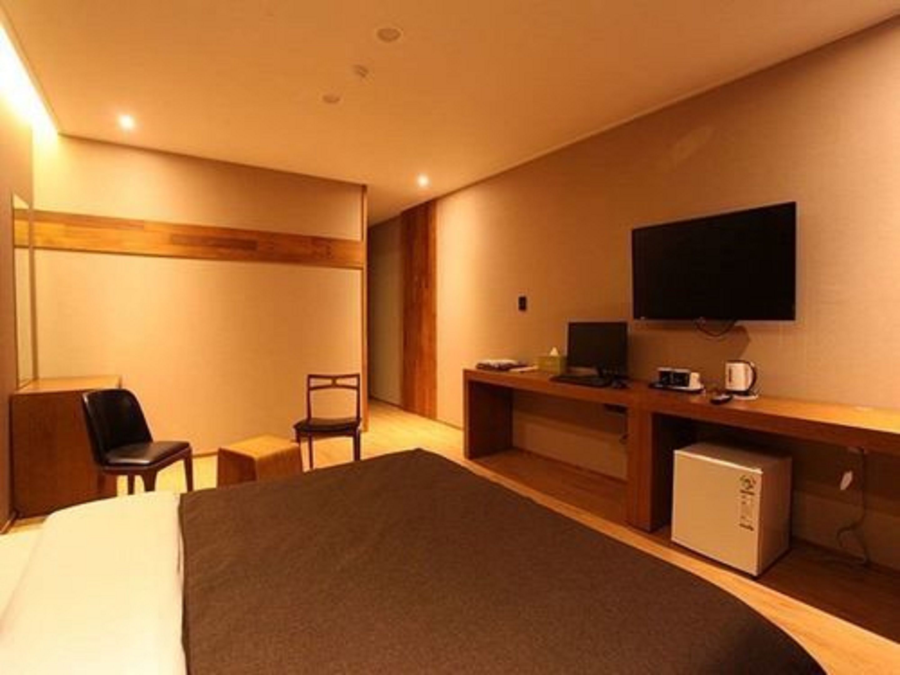 A hotel room with a bed, a desk with a computer, a chair, a TV mounted on the wall, and a small refrigerator.