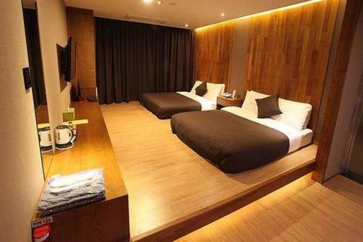 A modern hotel room with two beds, wooden flooring, a flat-screen TV, and a small table with a lamp.
