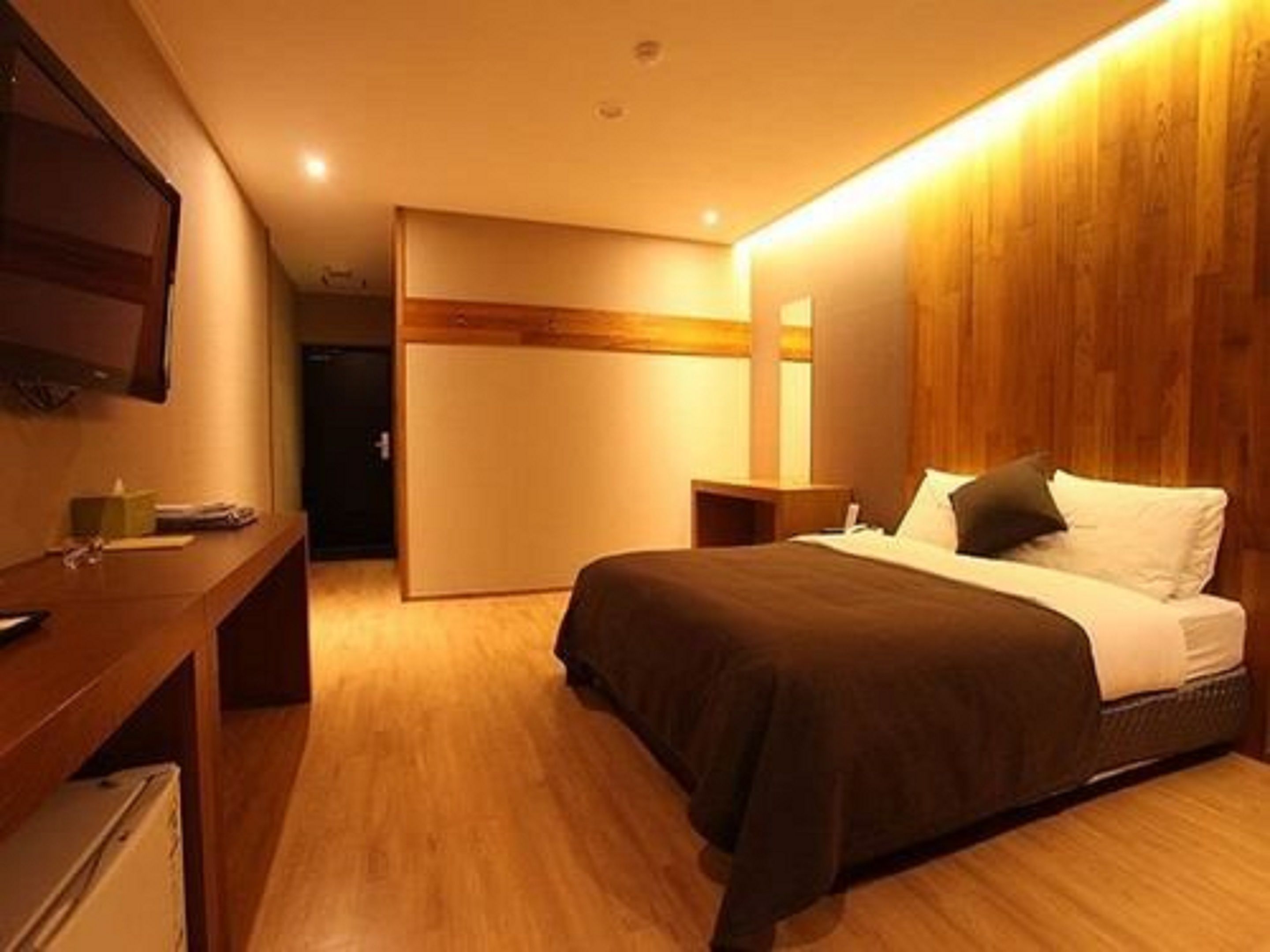 A modern hotel room with a wooden floor, a bed with a dark brown bedspread, a bedside table, and a flat-screen TV mounted on the wall.