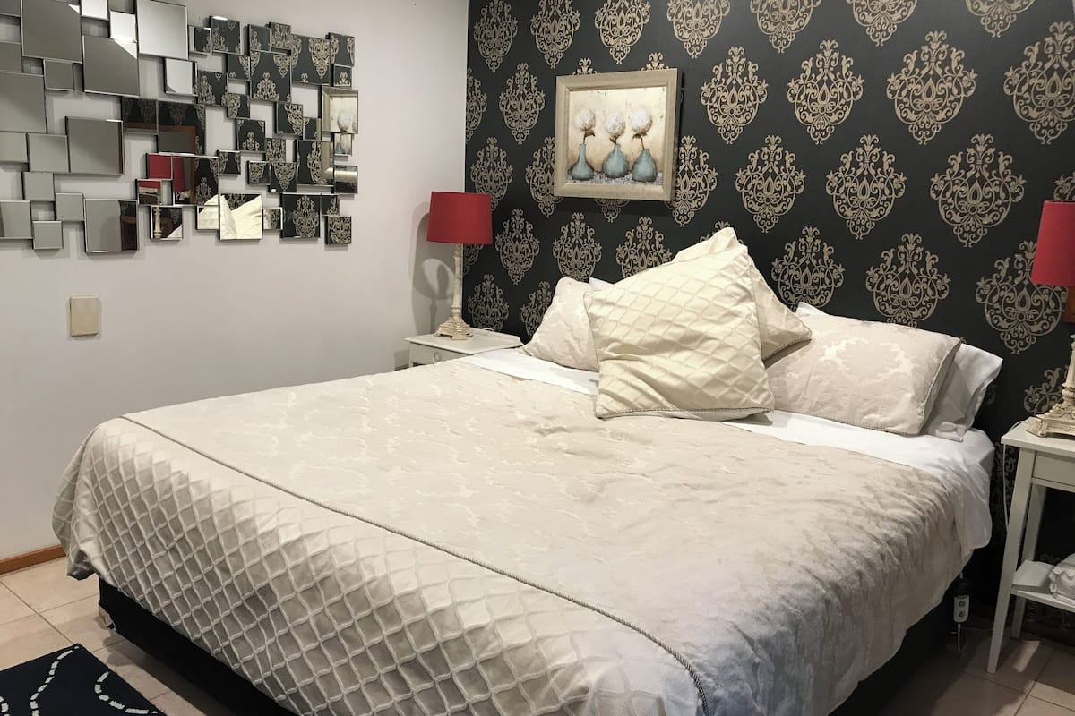 A neatly made bed with a quilted cover, a bedside table with a lamp, and a wall decorated with mirrors and a framed picture.